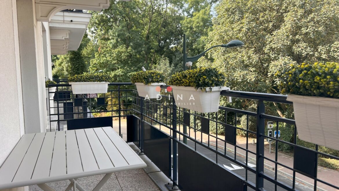 Balcon avec table et jardinières fleuries au-dessus d'une rue bordée d'arbres.