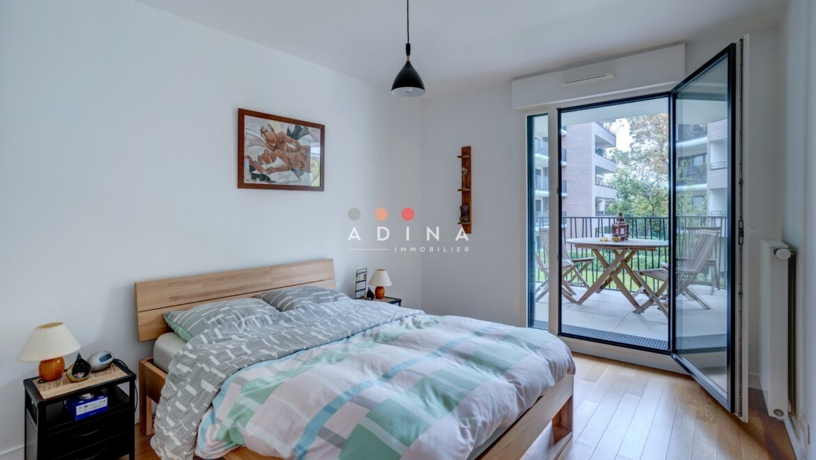 Chambre lumineuse avec balcon et vue sur des immeubles.