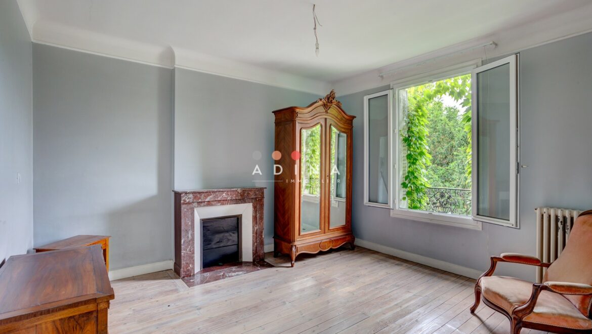 Salon avec cheminée et fenêtre ouverte, vue sur jardin verdoyant.