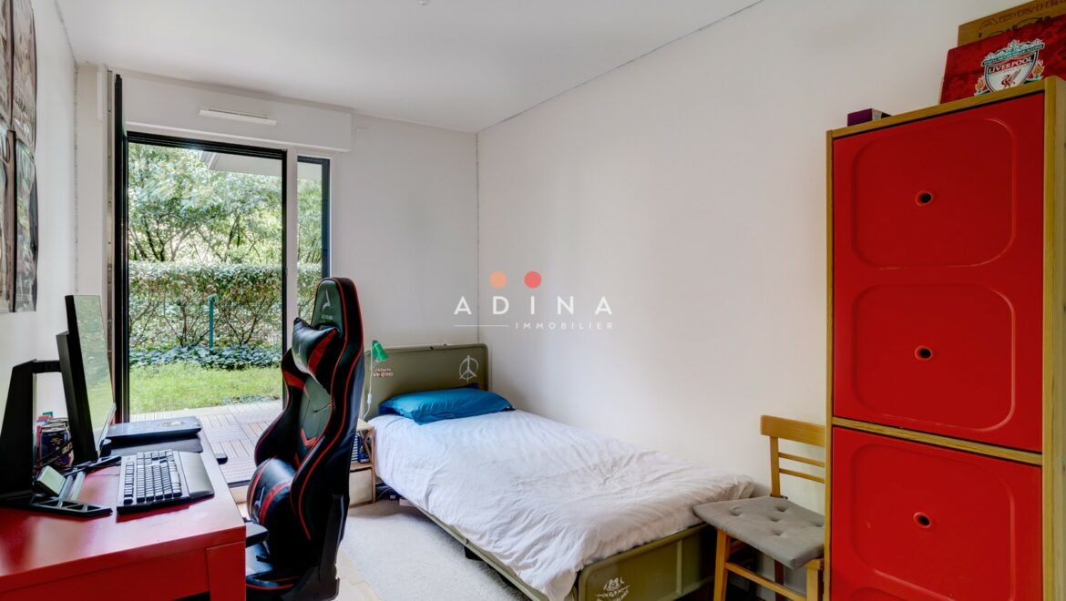 Chambre avec bureau, lit et armoire rouge ouvrant sur un jardin.